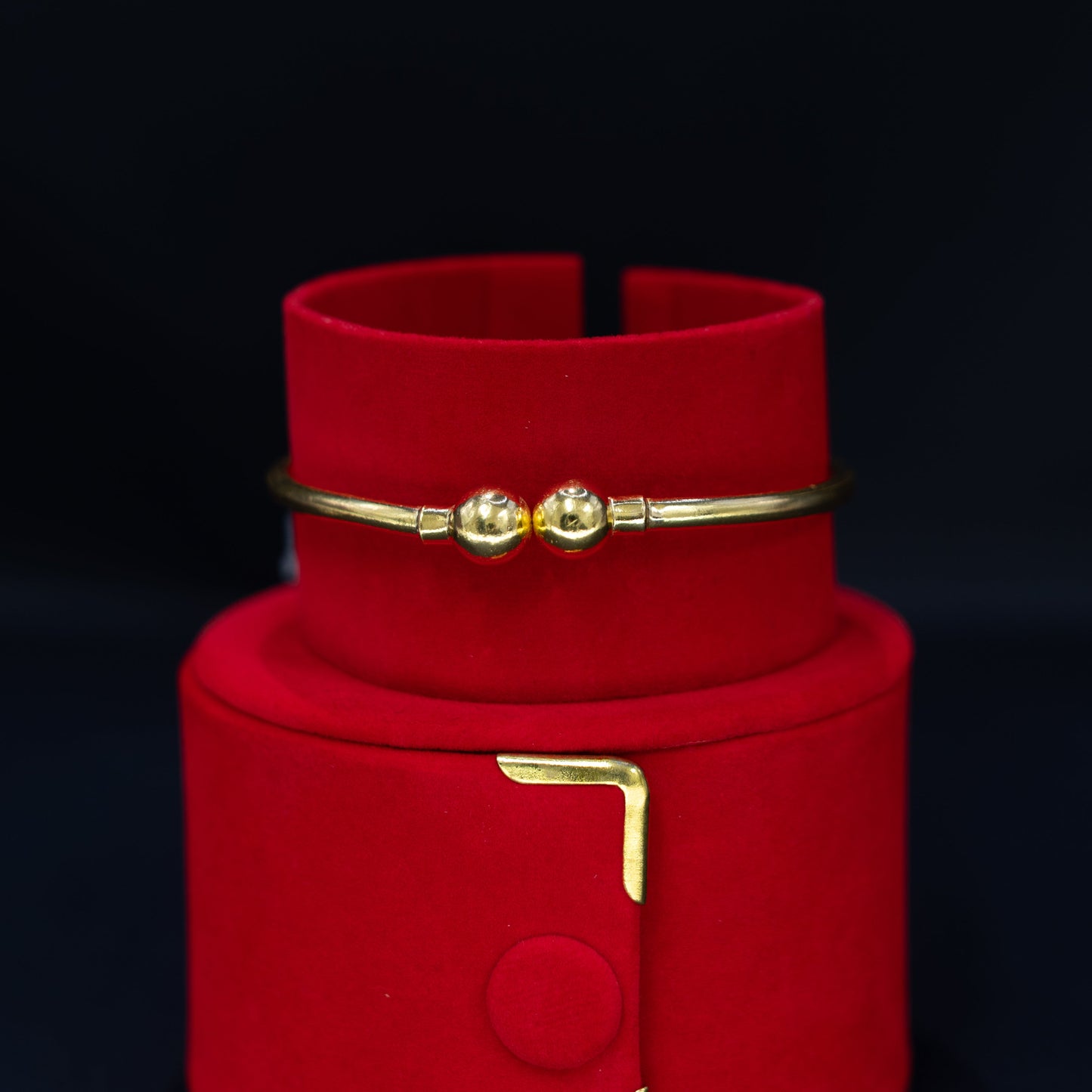 A gold bangle bracelet with two small bead details, displayed on a red velvet jewelry stand against a dark background.