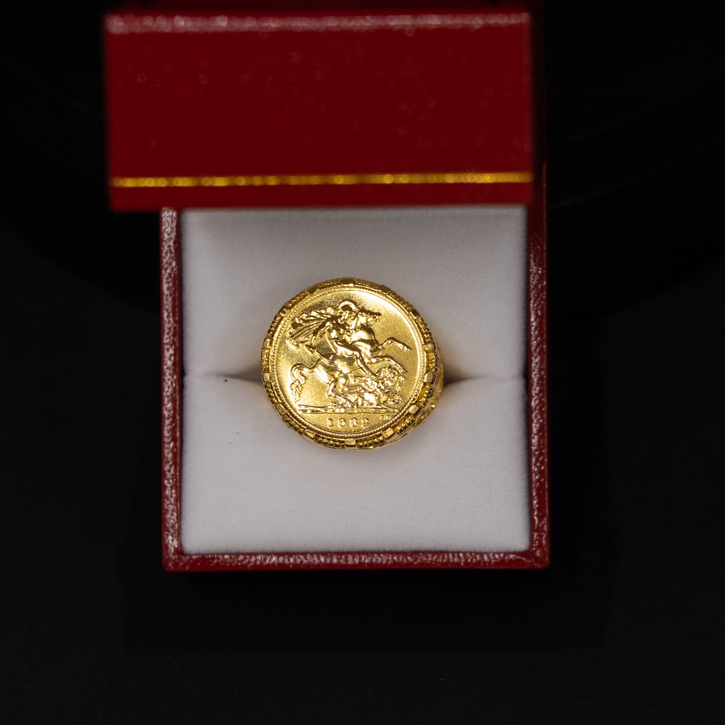 A round gold coin or medallion displayed inside an open red jewelry box with a white insert, set against a black background.