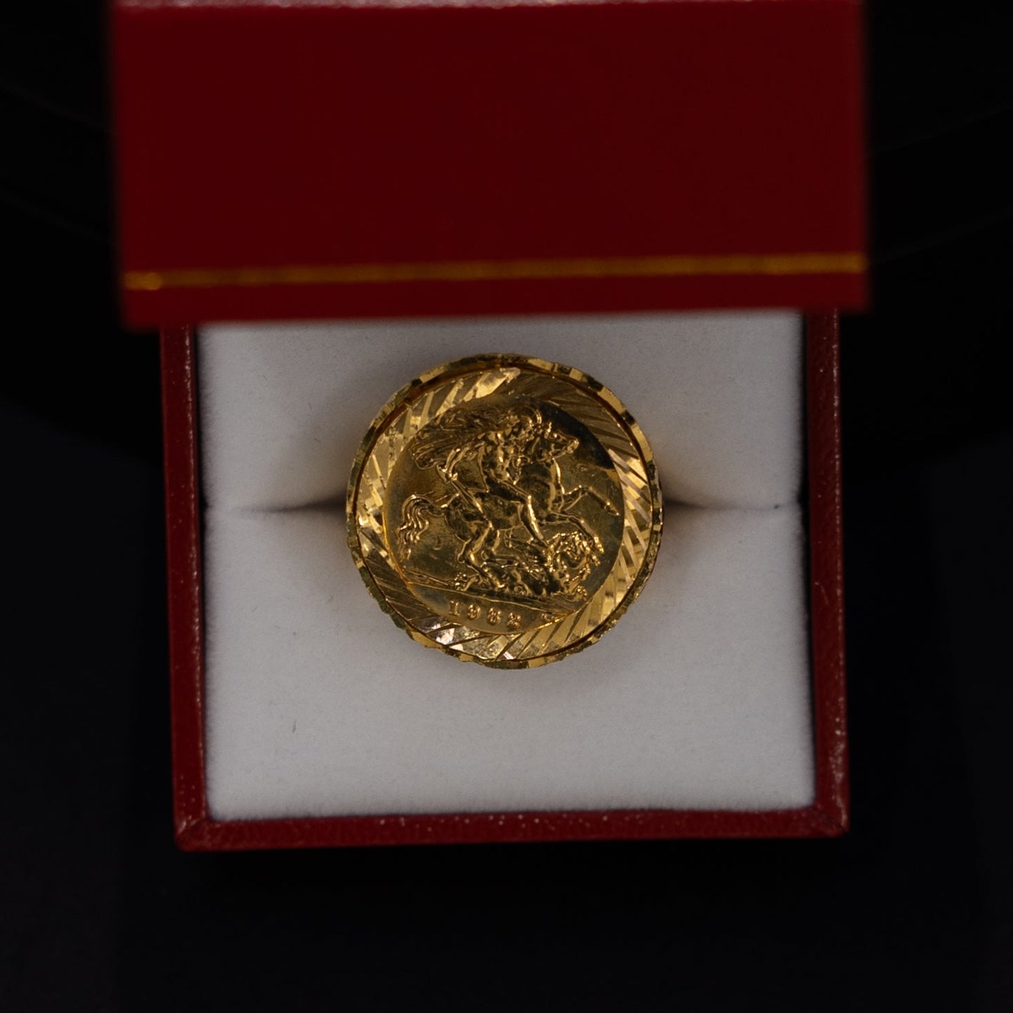 A small gold coin displayed inside an open red presentation box with a white insert, set against a dark black background.