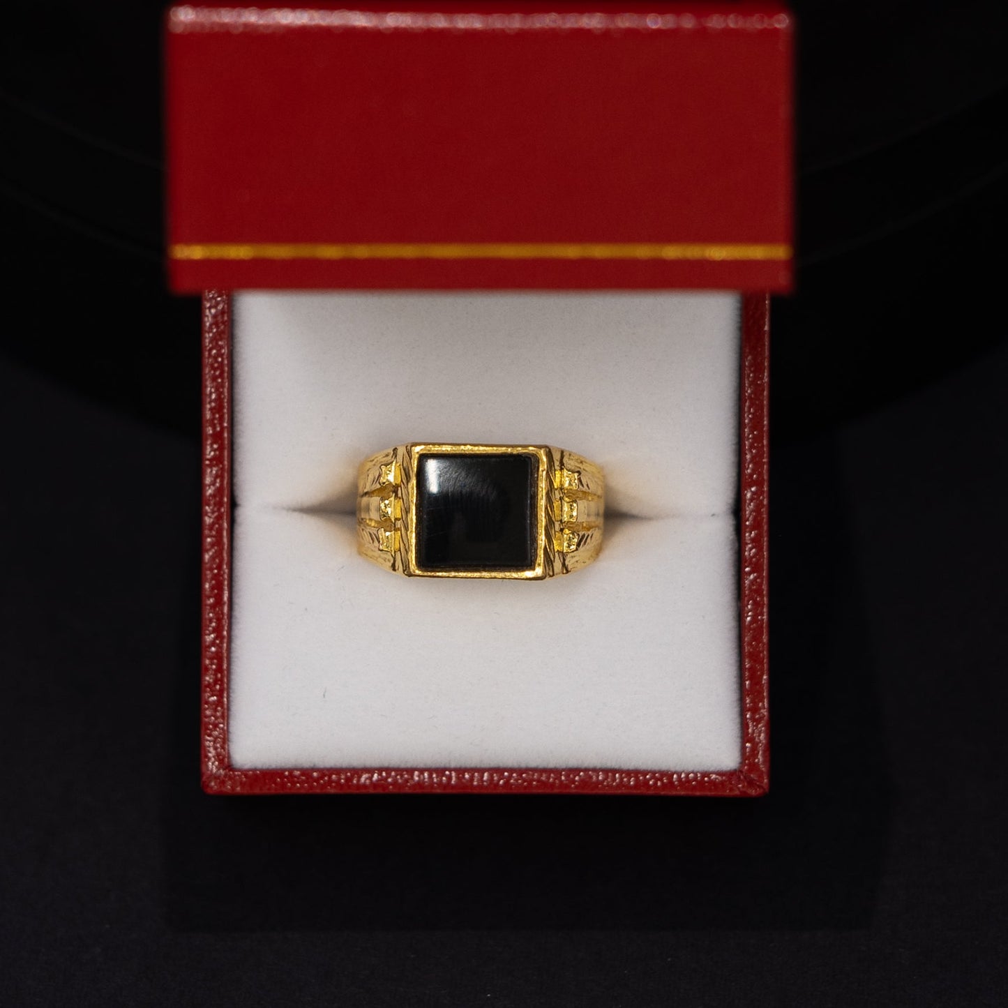 A gold signet ring with a square black stone, displayed inside an open red jewelry box on a black background.
