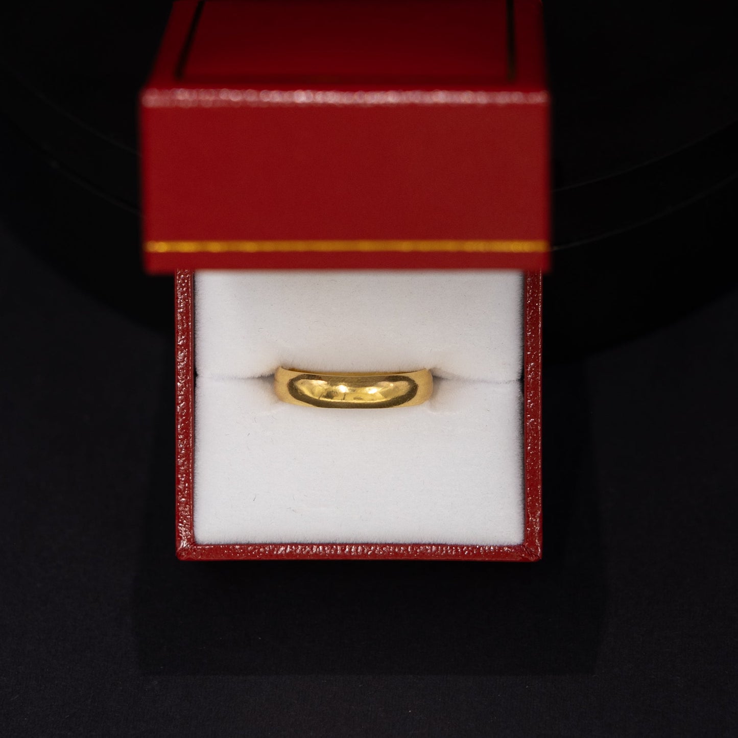 A gold band ring displayed inside an open red jewelry box with a white interior, set against a dark black background.