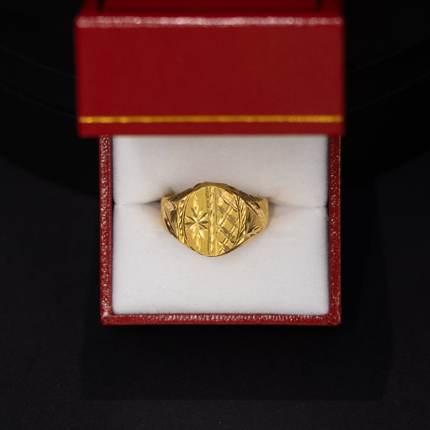 A gold signet ring displayed inside an open red jewelry box with a white insert, set against a dark black background.