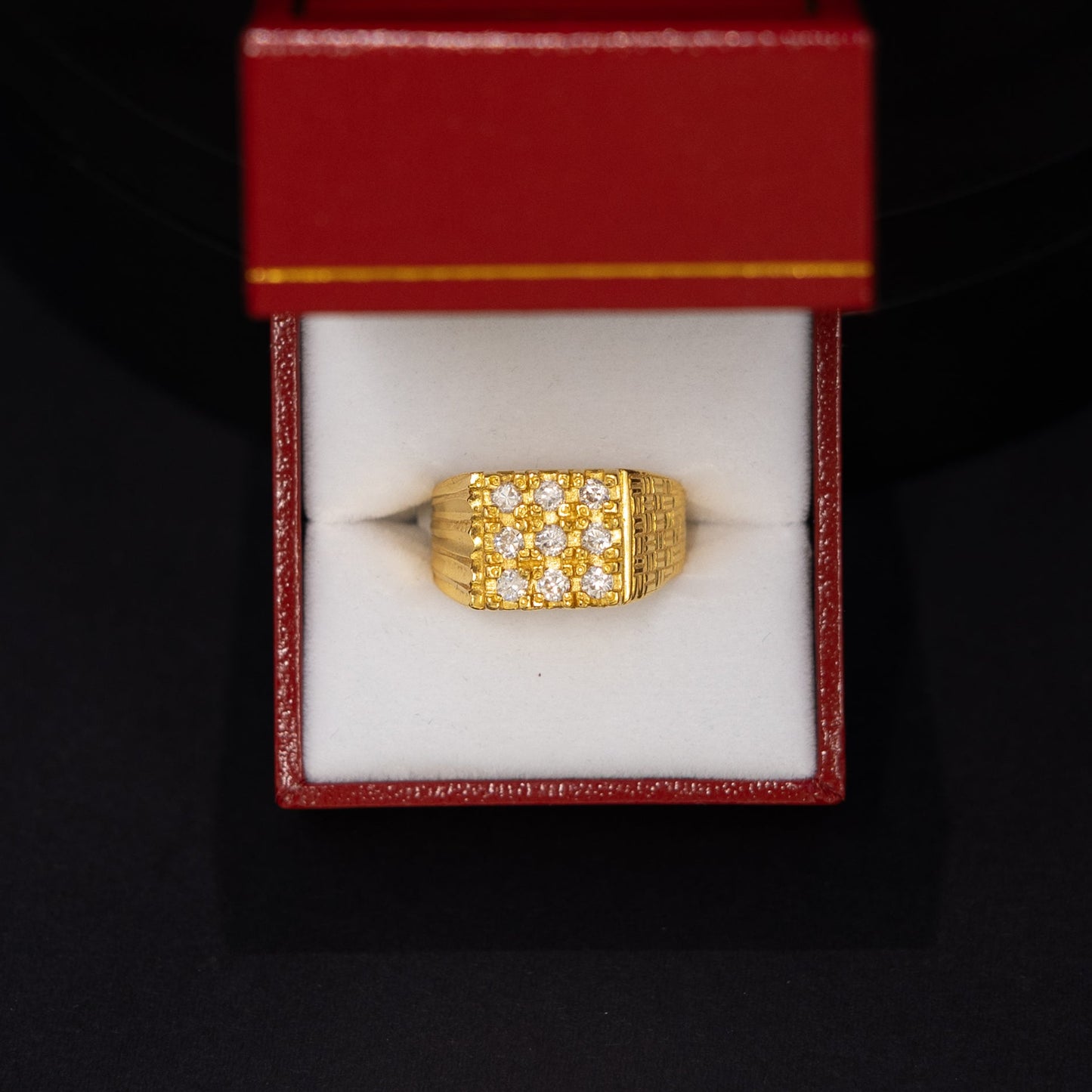 A gold ring with a textured, grid-like pattern, presented inside an open red jewelry box with a white insert, set against a dark black background.