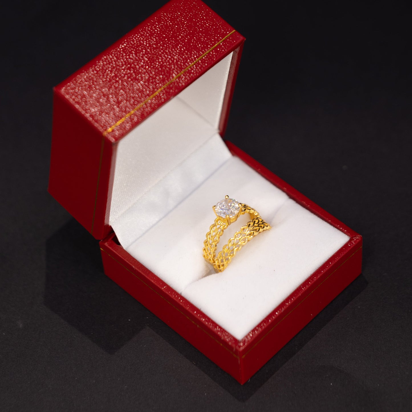 A gold ring with a clear center stone displayed inside an open red jewelry box with a white velvet lining, set against a dark black background.