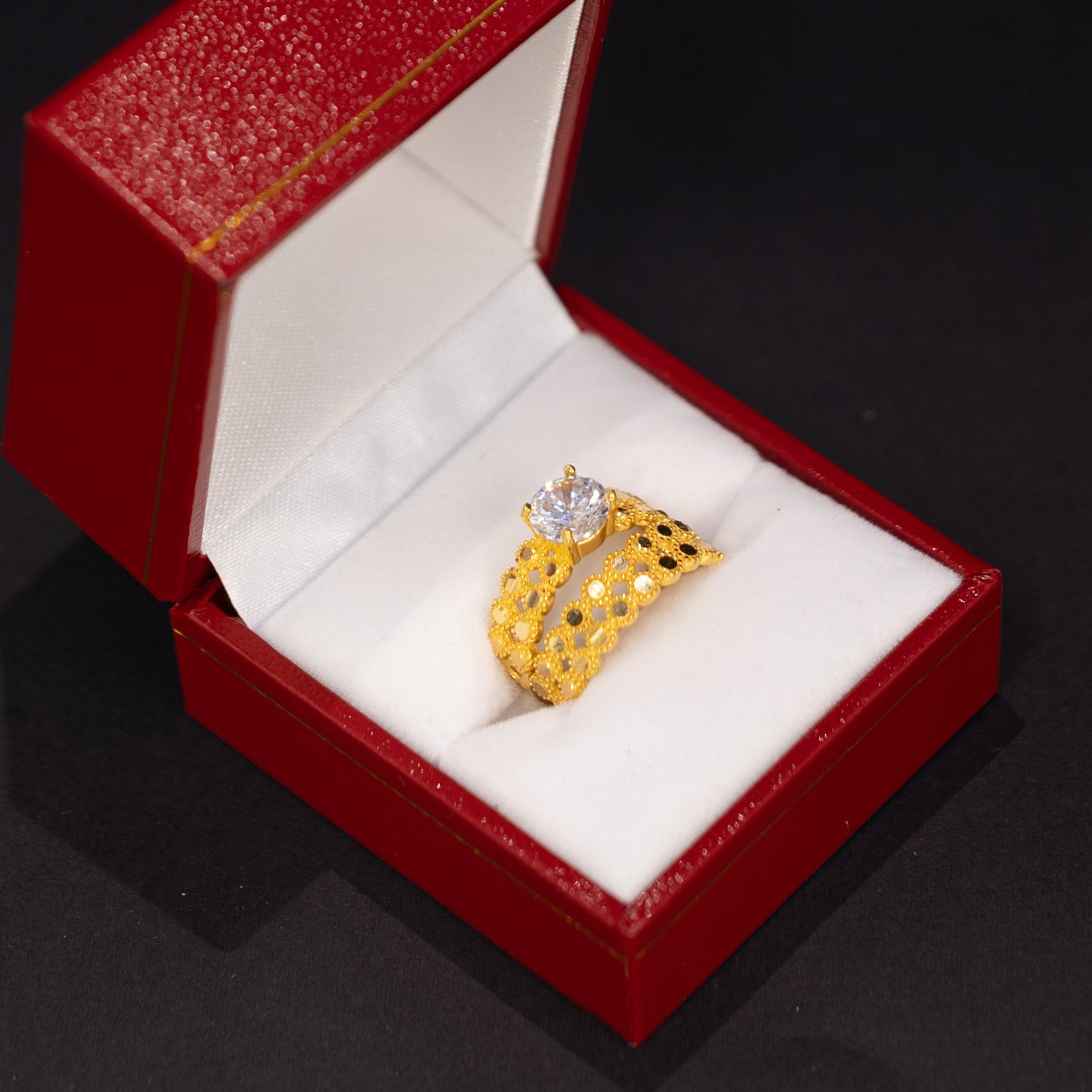A yellow gold ring with a clear central gemstone, displayed inside an open red jewelry box with a white cushion on a dark background.