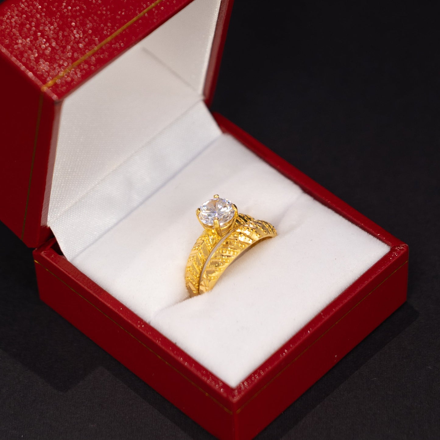 A gold ring with a clear center stone displayed inside an open red jewelry box with a white velvet lining, set against a dark black background.