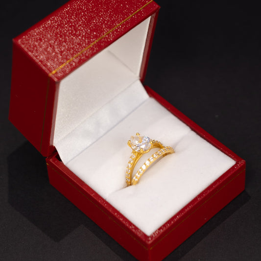 A gold ring with a clear central gemstone and small side stones, displayed inside an open red jewelry box with a white interior on a dark background.