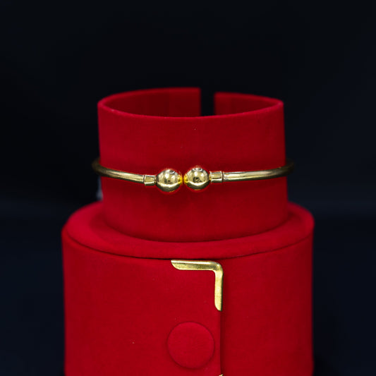 A gold bangle bracelet with two small bead details, displayed on a red velvet jewelry stand against a dark background.