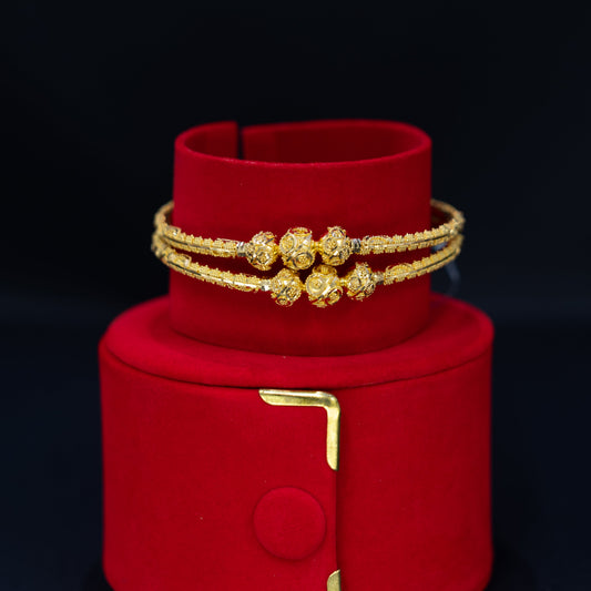 A pair of gold double bangles with textured floral motifs and a central gold bead, displayed on a red velvet jewelry stand against a black background.