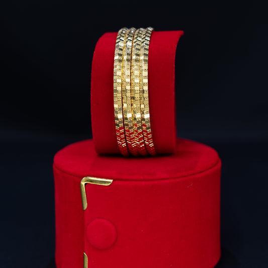 A set of gold-toned textured bangles displayed on a red velvet cylindrical stand, which is placed on top of a matching red velvet cylindrical box against a black background.