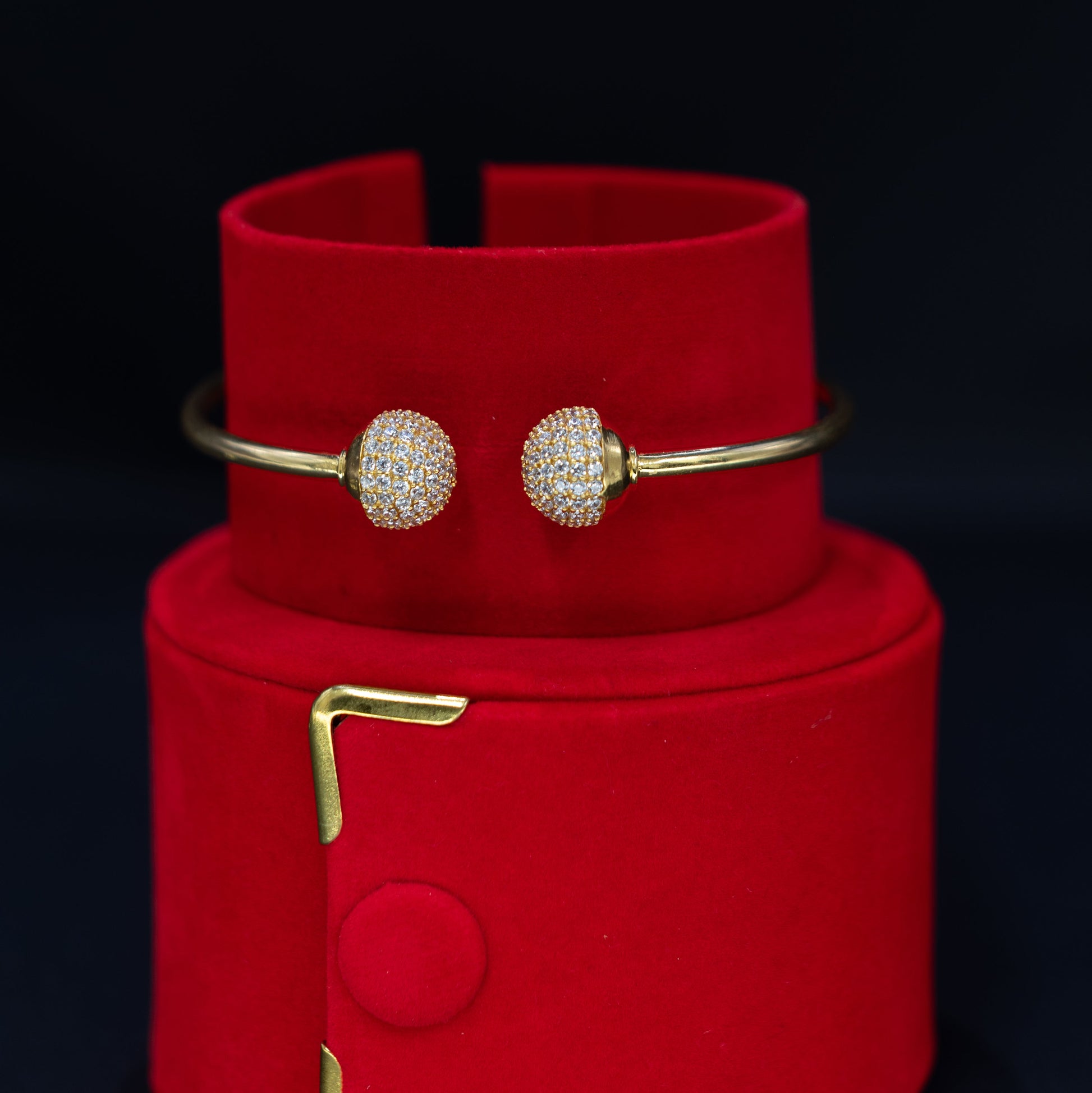 A pair of gold bangle bracelets with round pavé-set crystal clusters, displayed on a red velvet jewelry stand against a black background.