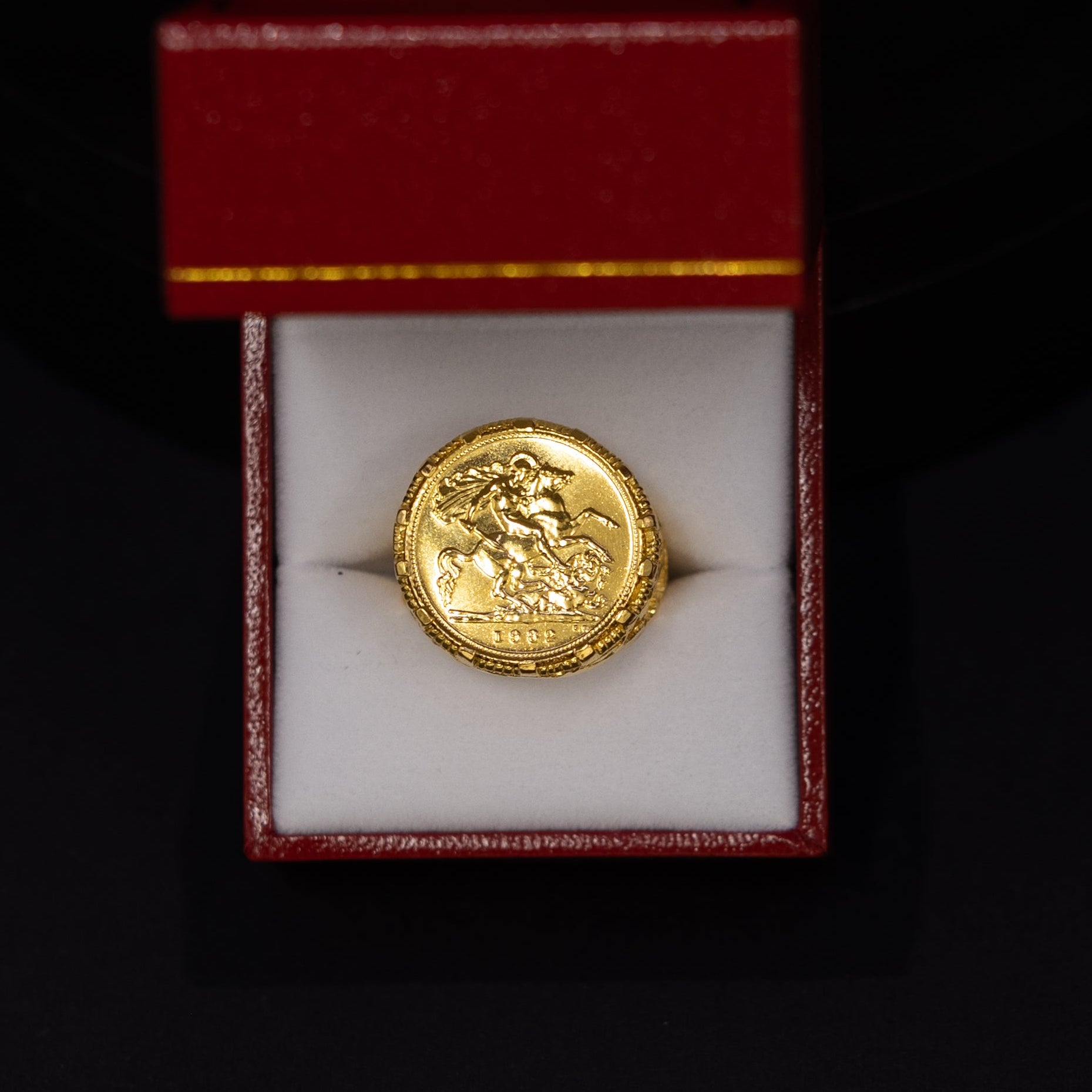 A round gold coin or medallion displayed inside an open red jewelry box with a white insert, set against a black background.