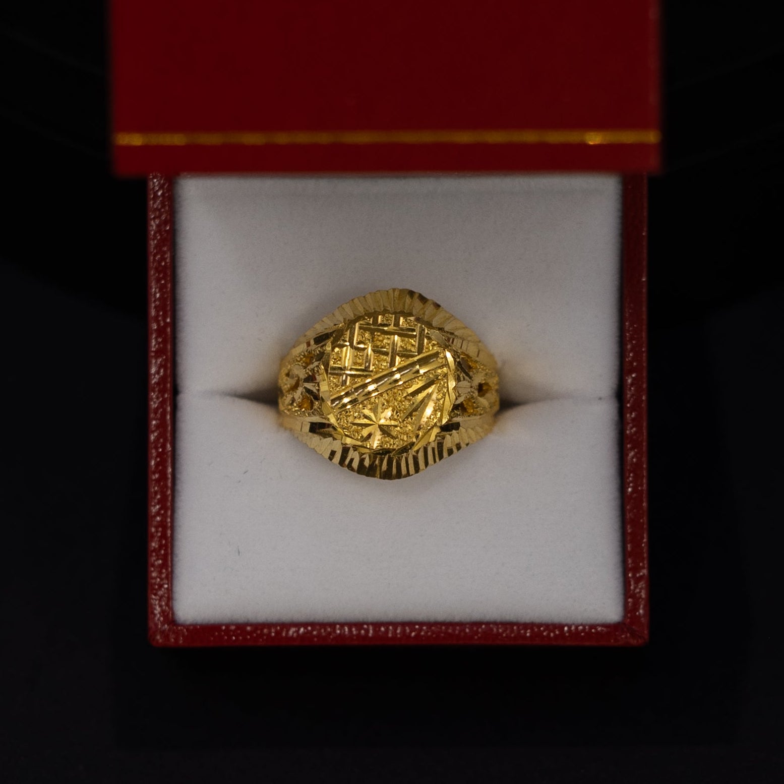A gold signet ring with an embossed crest, presented inside an open red jewelry box with a white velvet cushion against a black background.