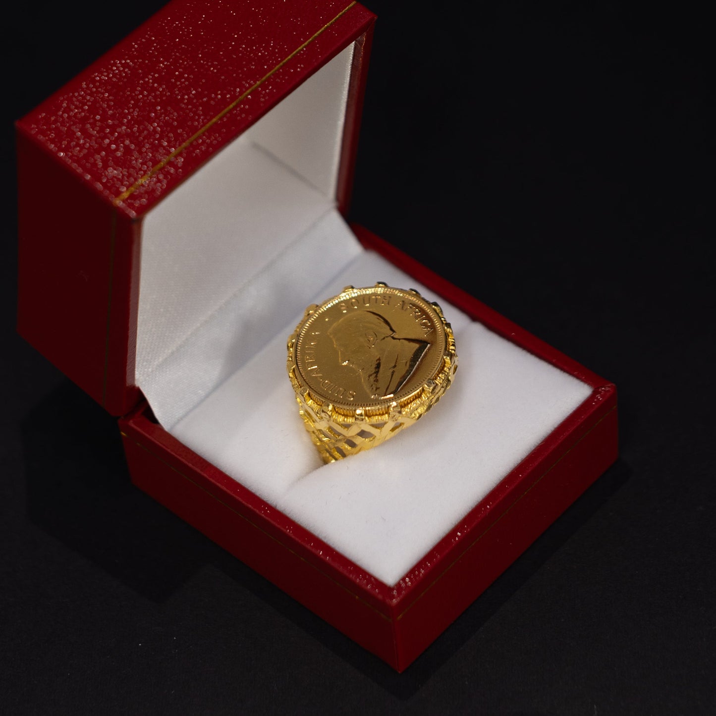 A gold ring displayed inside an open red presentation box with a white velvet insert, set against a dark black background.