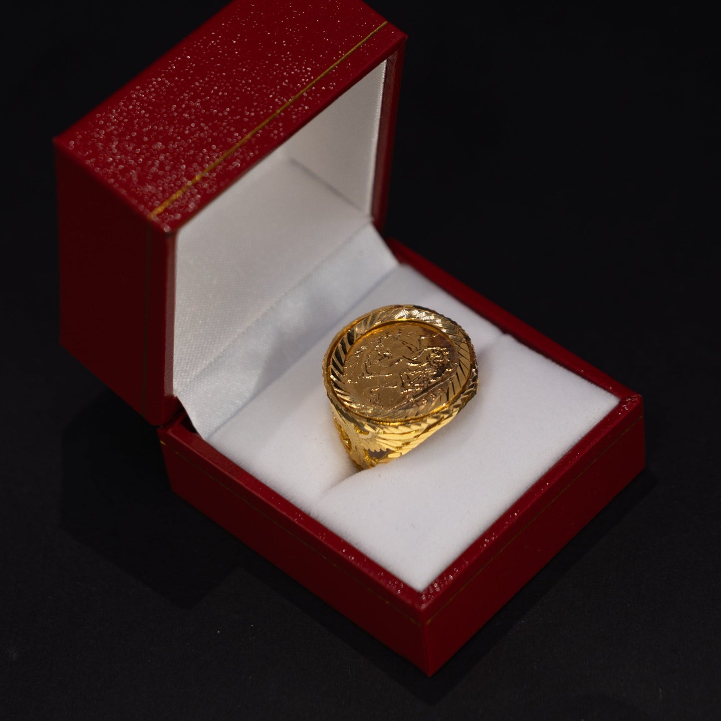 A gold signet ring with an embossed crest, displayed inside an open red velvet jewelry box with a white cushion on a black background.