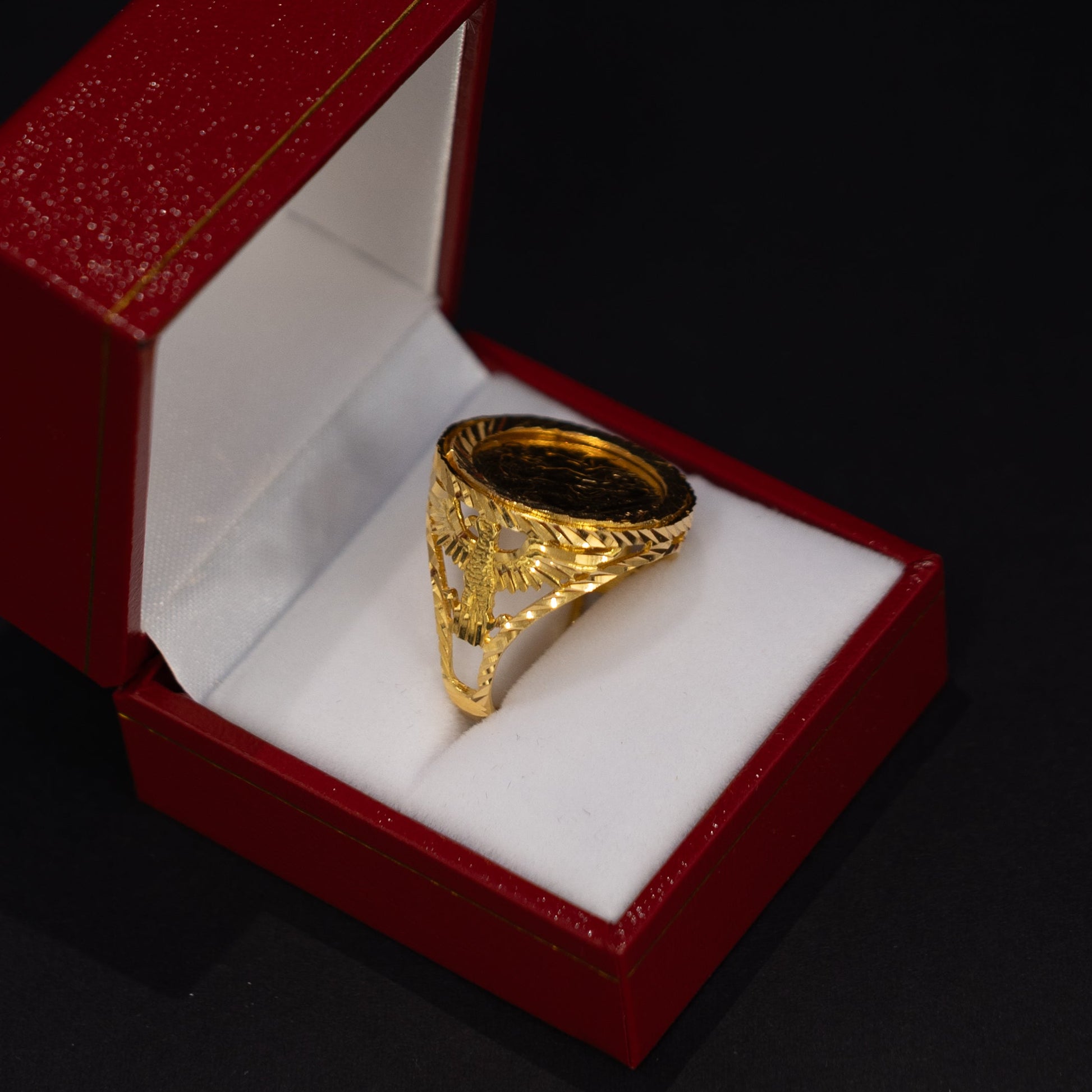 A gold signet ring with an engraved triangular face, displayed inside an open red velvet jewelry box with a white cushion on a black background.