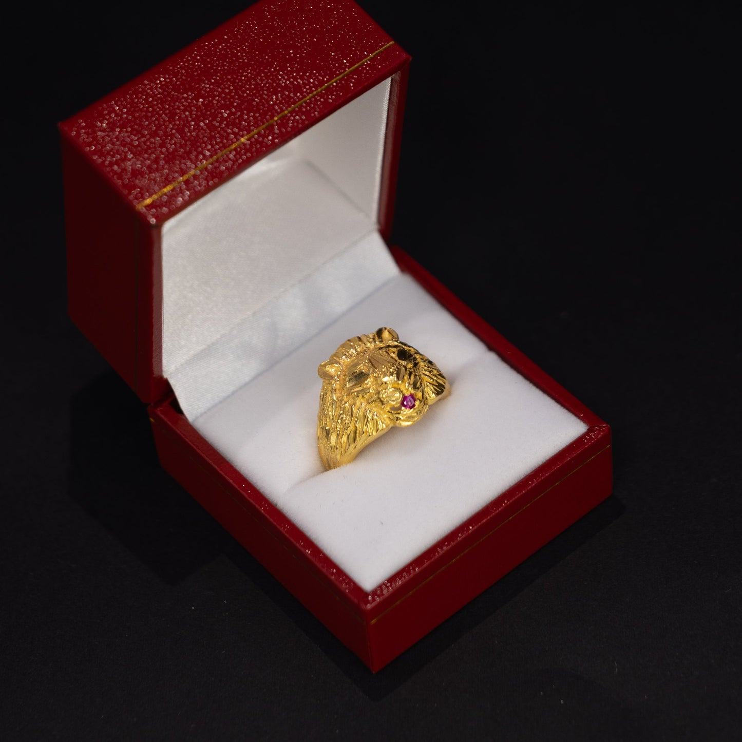 A gold ring with small red gemstone accents sits inside an open red jewelry box with a white cushion, set against a dark black background.