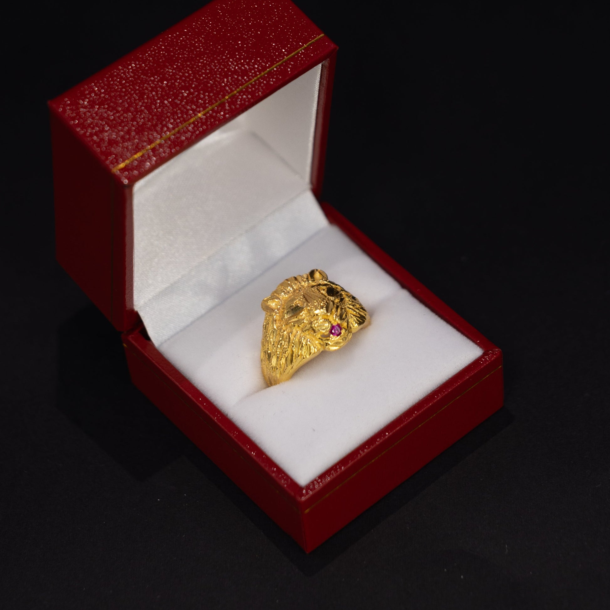 A gold ring with small red gemstone accents sits inside an open red jewelry box with a white cushion, set against a dark black background.