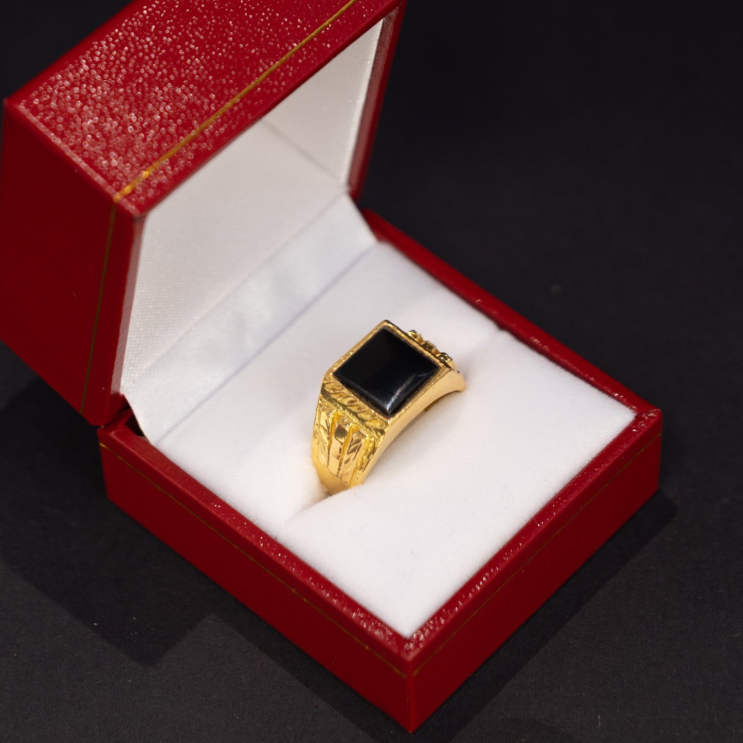 A gold signet-style ring with a rectangular black inlay, displayed inside an open red jewelry box with white cushioning on a dark background.