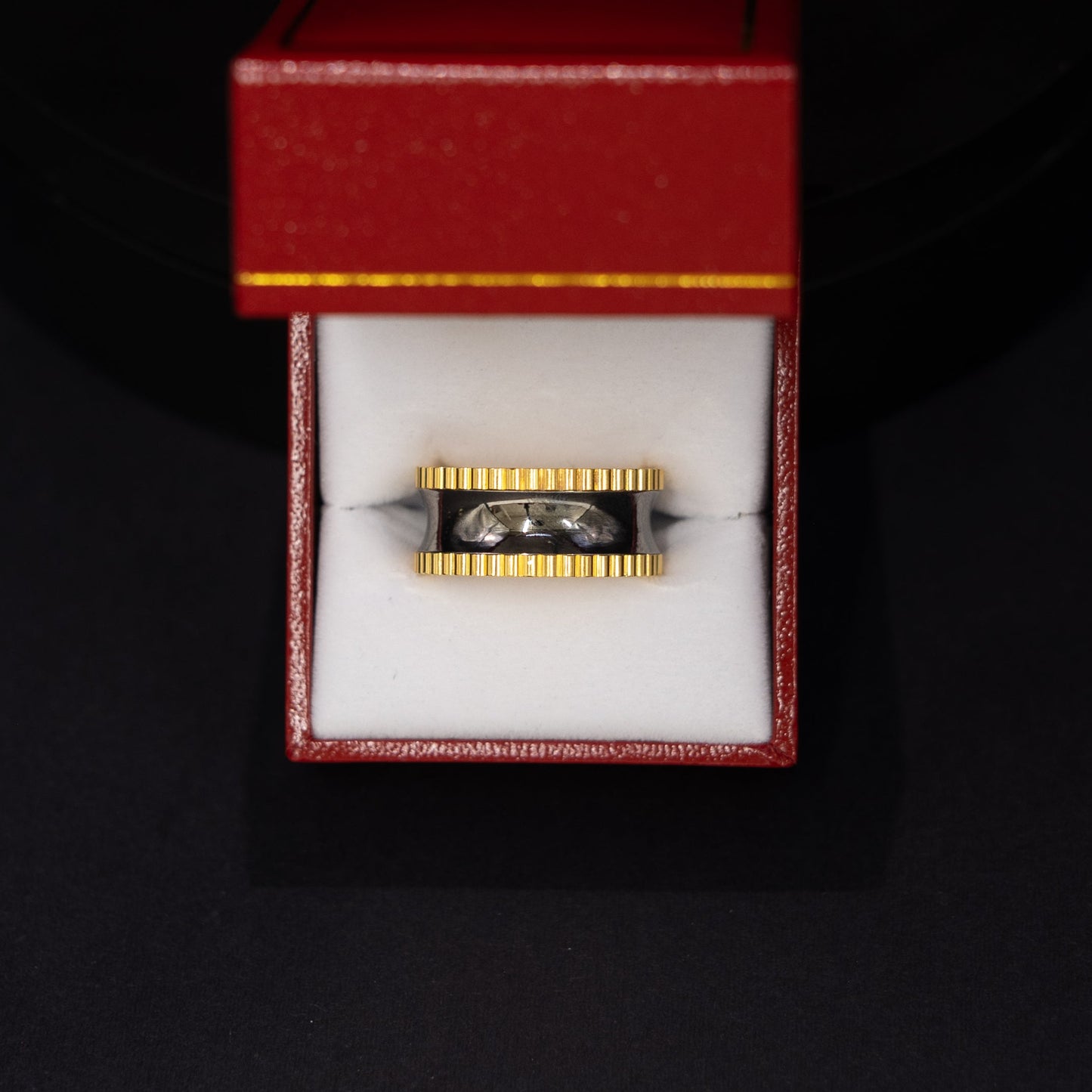A gold and black ring displayed inside an open red jewelry box with a white interior, set against a dark black background.