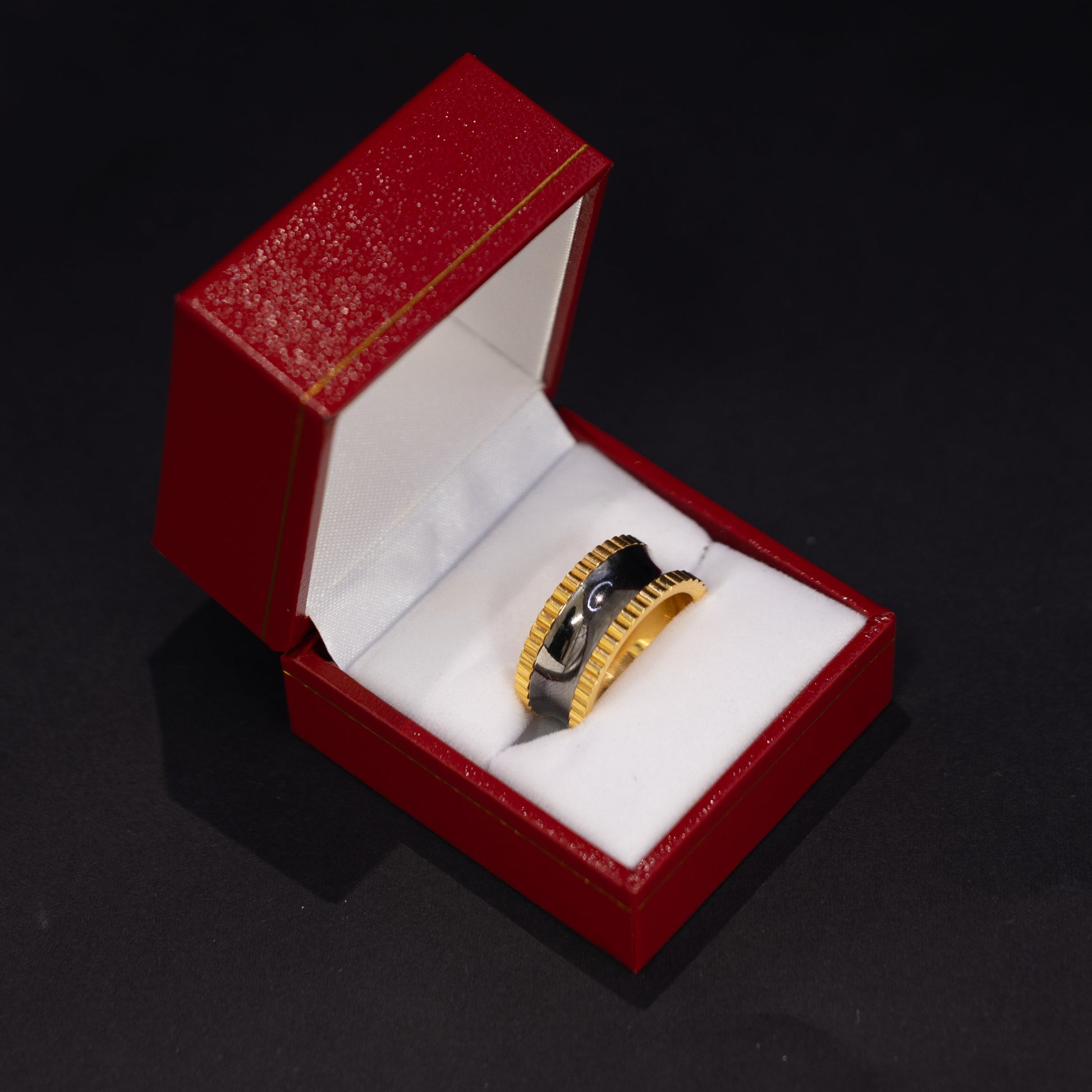 A gold ring with a black center and textured detailing, displayed inside an open red jewelry box with a white cushion on a dark background.