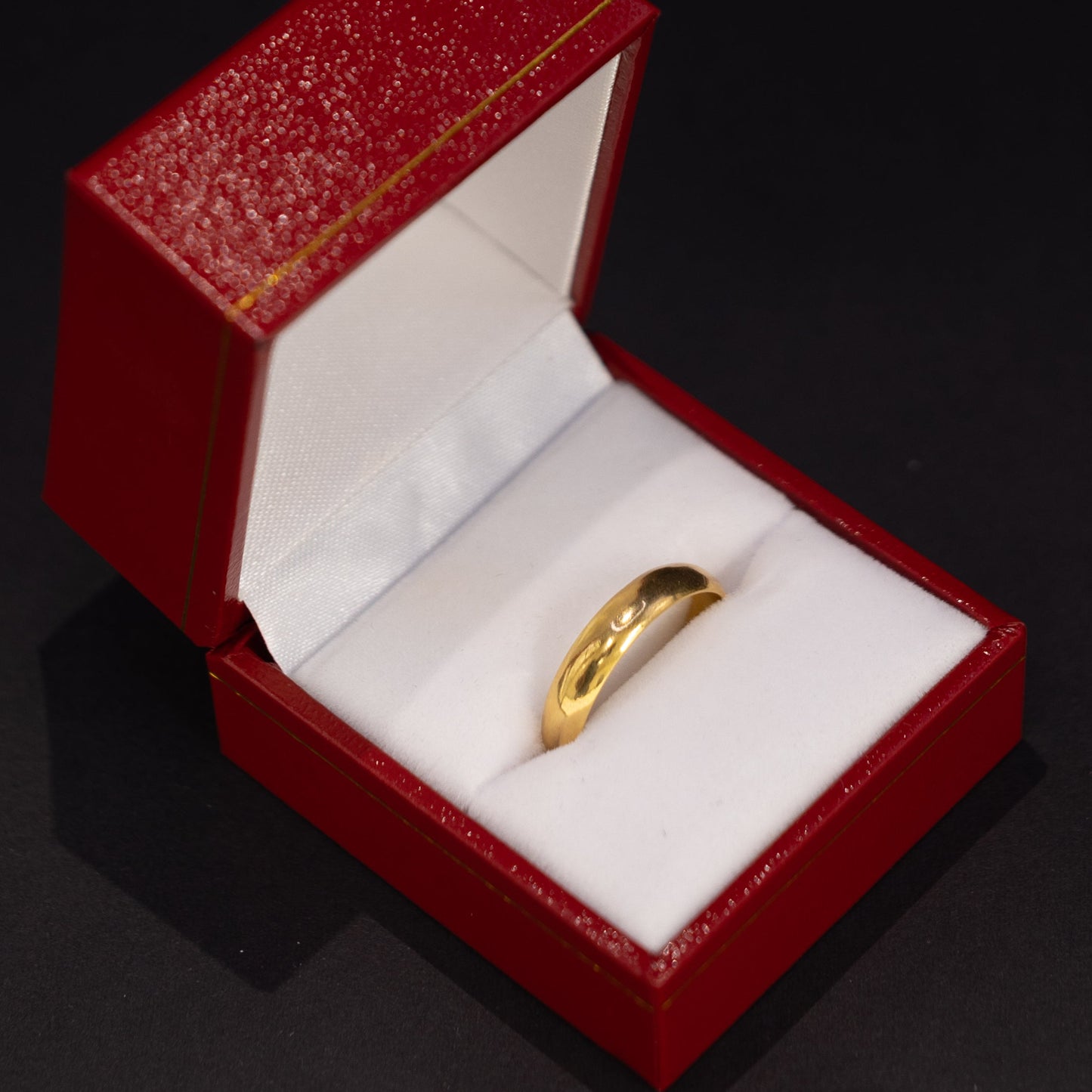 A gold band ring displayed inside an open red jewelry box with a white cushion, set against a dark black background.