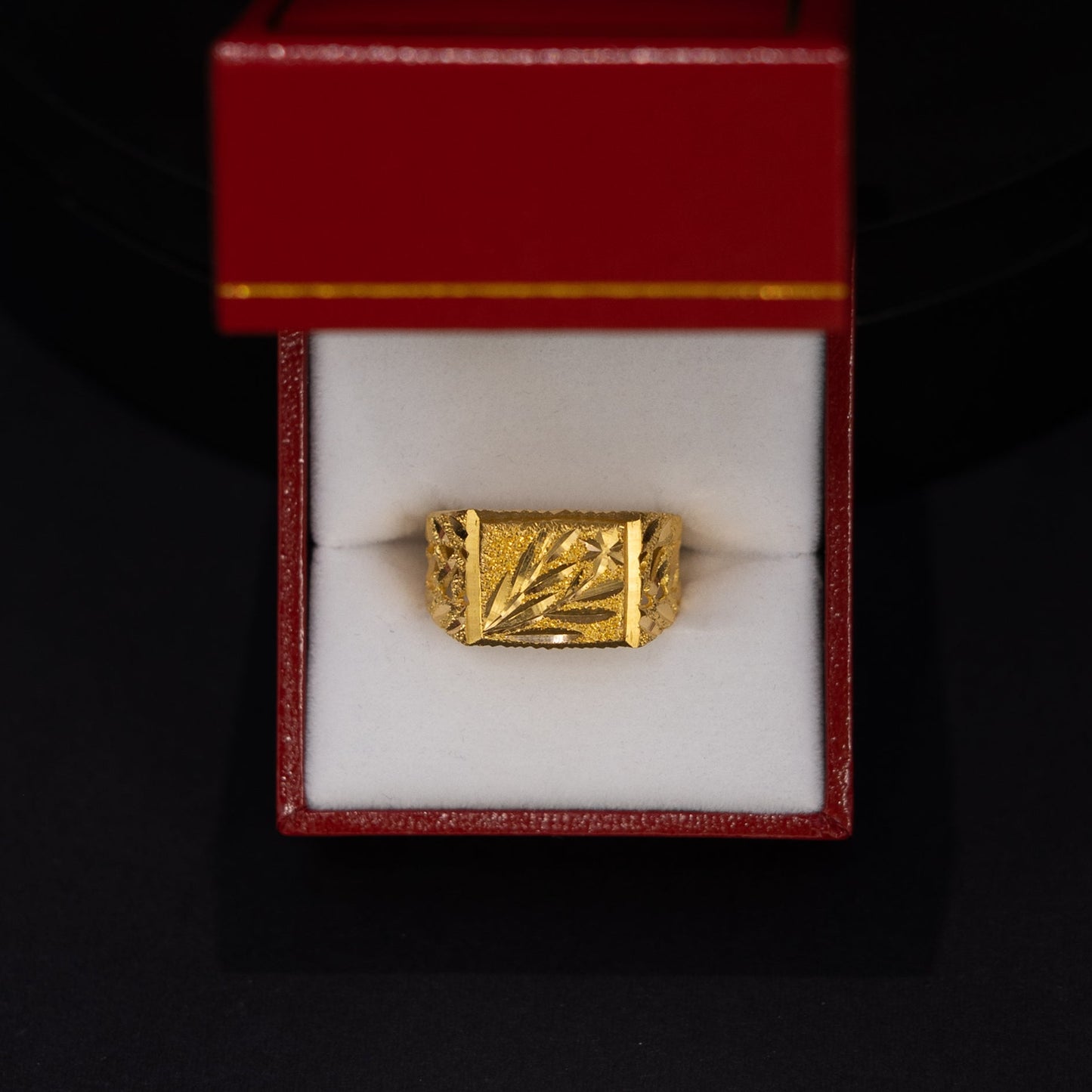 A gold ring with an engraved geometric pattern, presented in an open red jewelry box with a white interior, set against a dark black background.