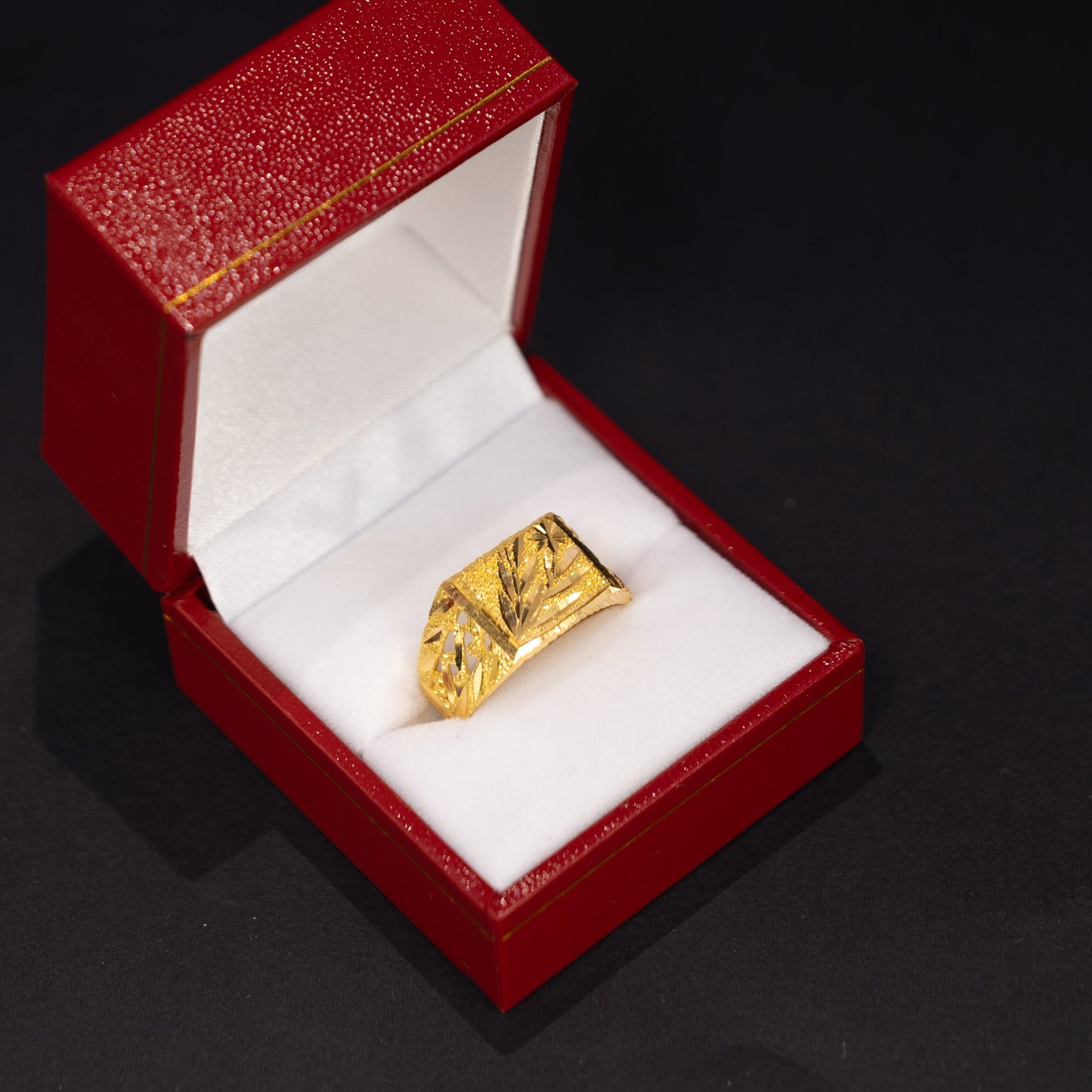 A gold ring with a textured, geometric design displayed inside an open red jewelry box with a white interior, set against a dark black background.