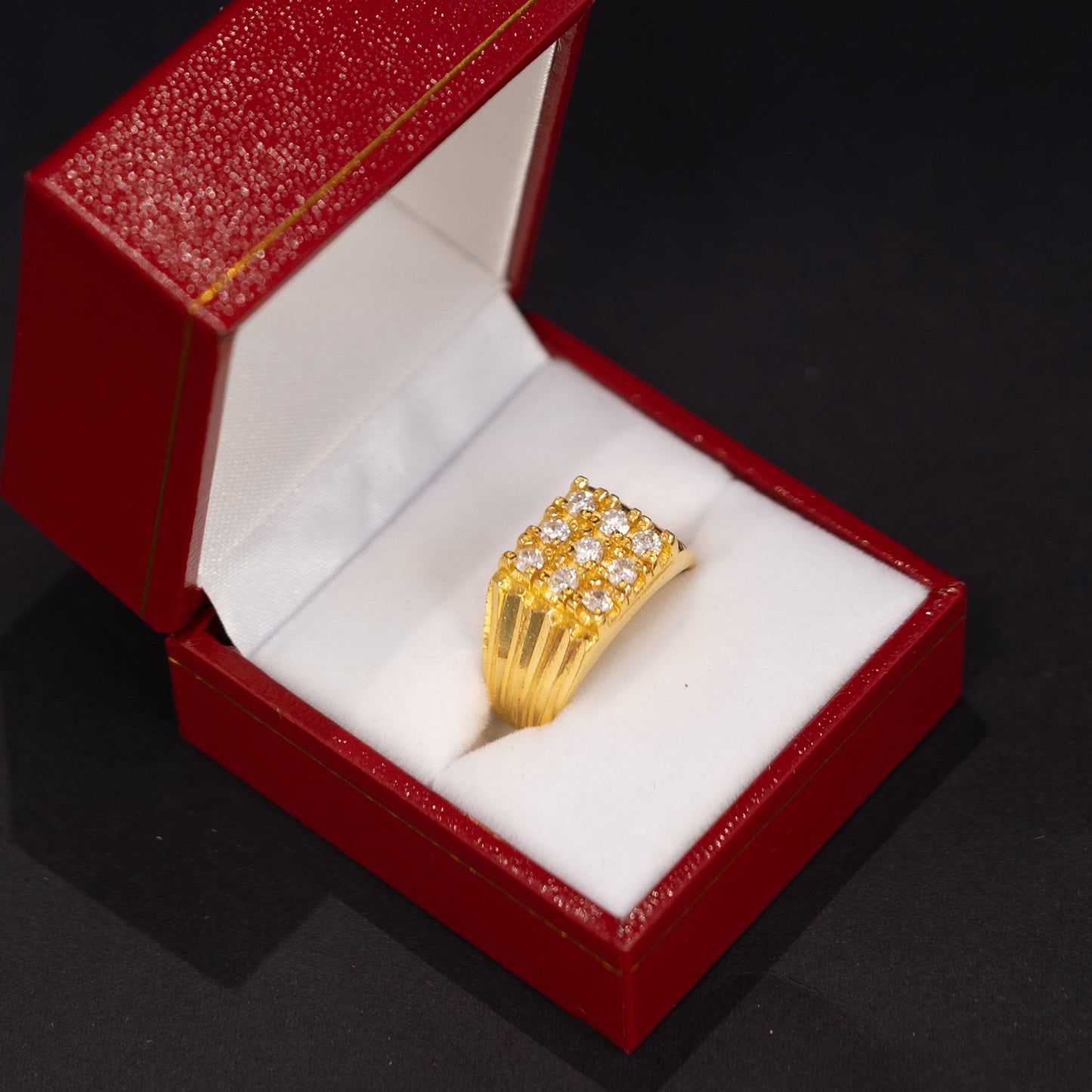 A gold ring with a textured, multi-row band displayed inside an open red jewelry box with a white cushion, set against a dark black background.
