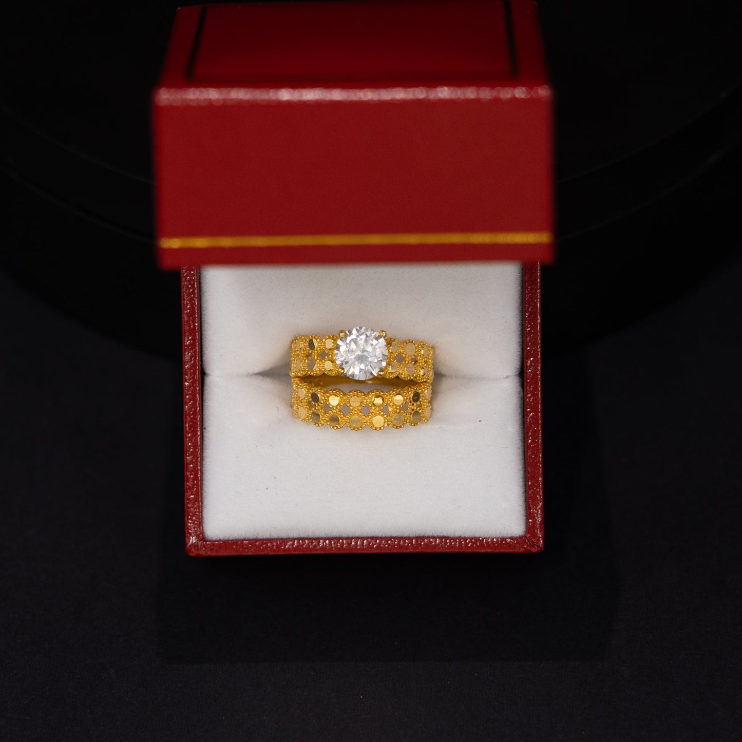 A yellow gold ring with a single round-cut clear gemstone, presented in an open red jewelry box with a white interior, set against a dark black background.