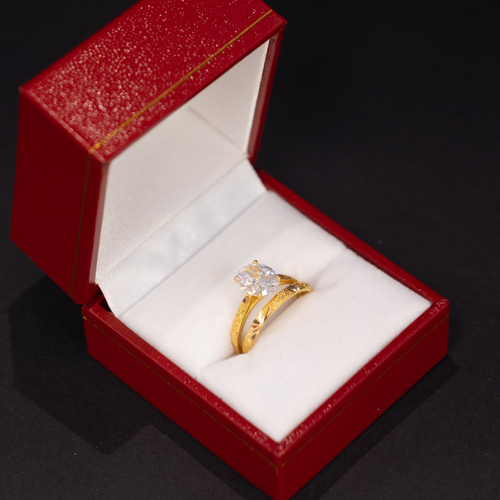 A gold ring with a clear center stone displayed in an open red jewelry box with a white cushion, set against a dark background.