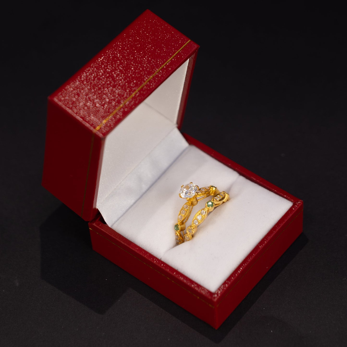 A gold ring with a clear gemstone displayed inside an open red jewelry box with a white velvet lining, set against a dark black background.