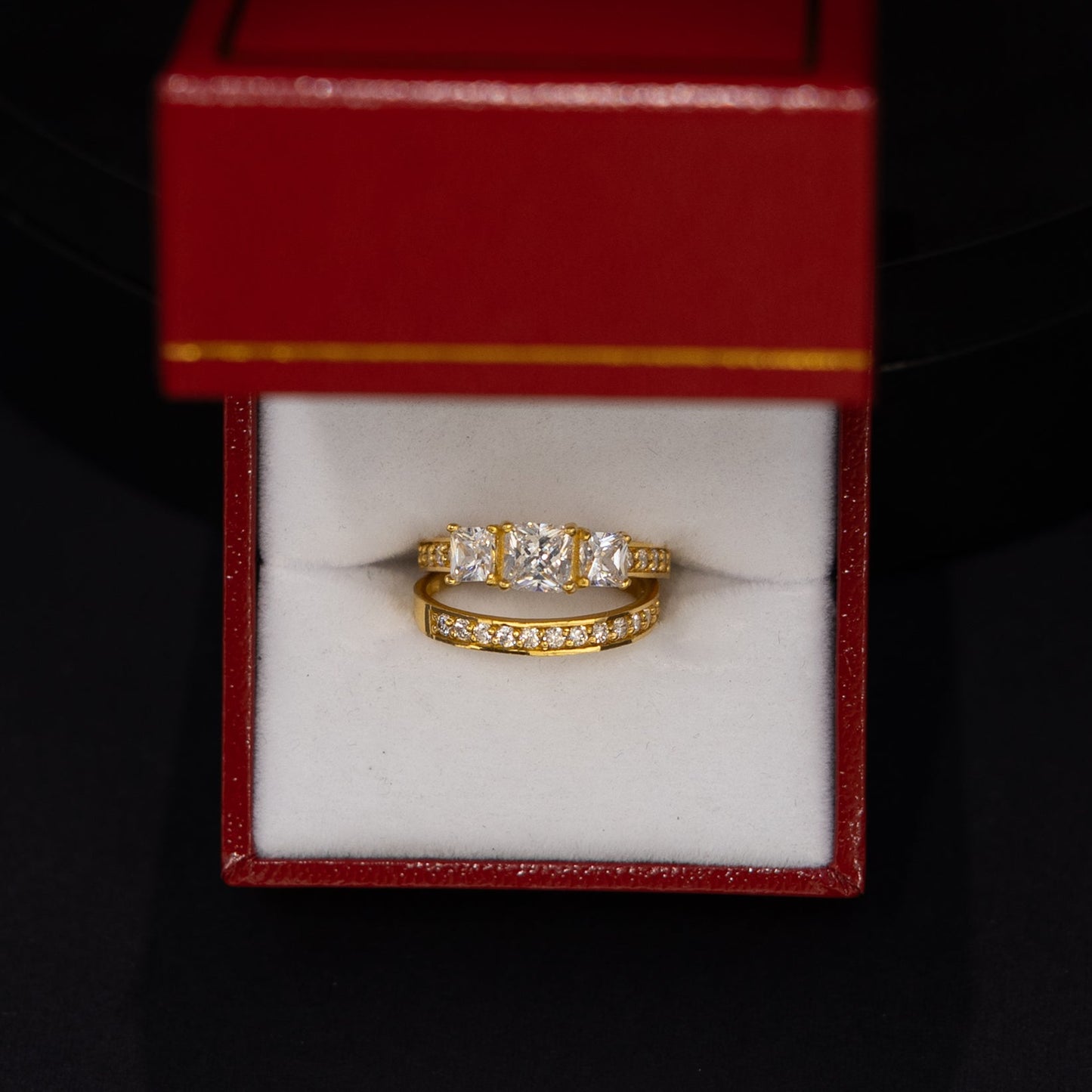 A yellow gold ring with small clear gemstones set in a white-lined red jewelry box, photographed against a dark black background.