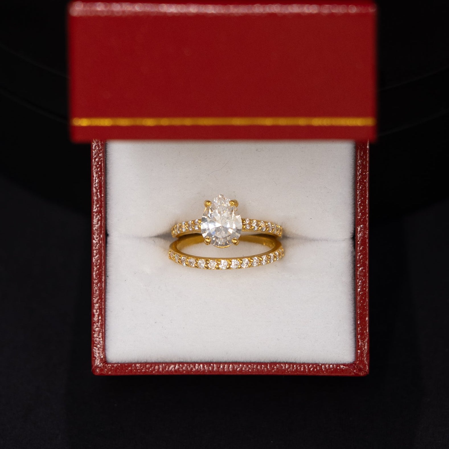 A gold ring with a central clear gemstone and a small band of clear stones, displayed inside an open red jewelry box with a white cushion, set against a dark black background.