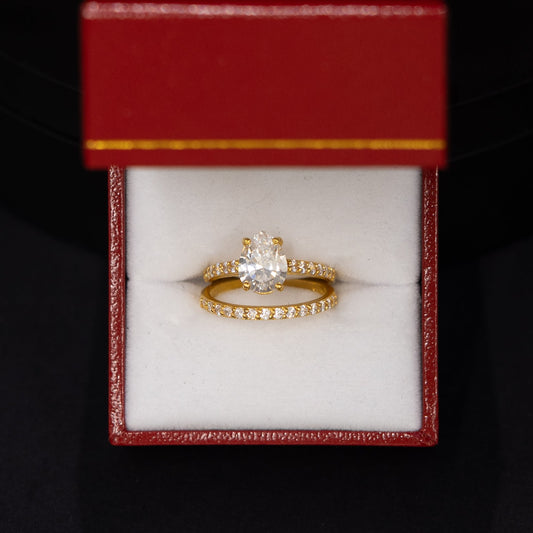 A gold ring with a central clear gemstone and a small band of clear stones, displayed inside an open red jewelry box with a white cushion, set against a dark black background.
