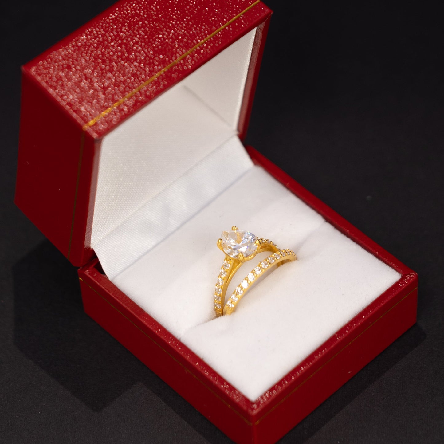 A gold ring with a clear central gemstone and small side stones, displayed inside an open red jewelry box with a white interior on a dark background.