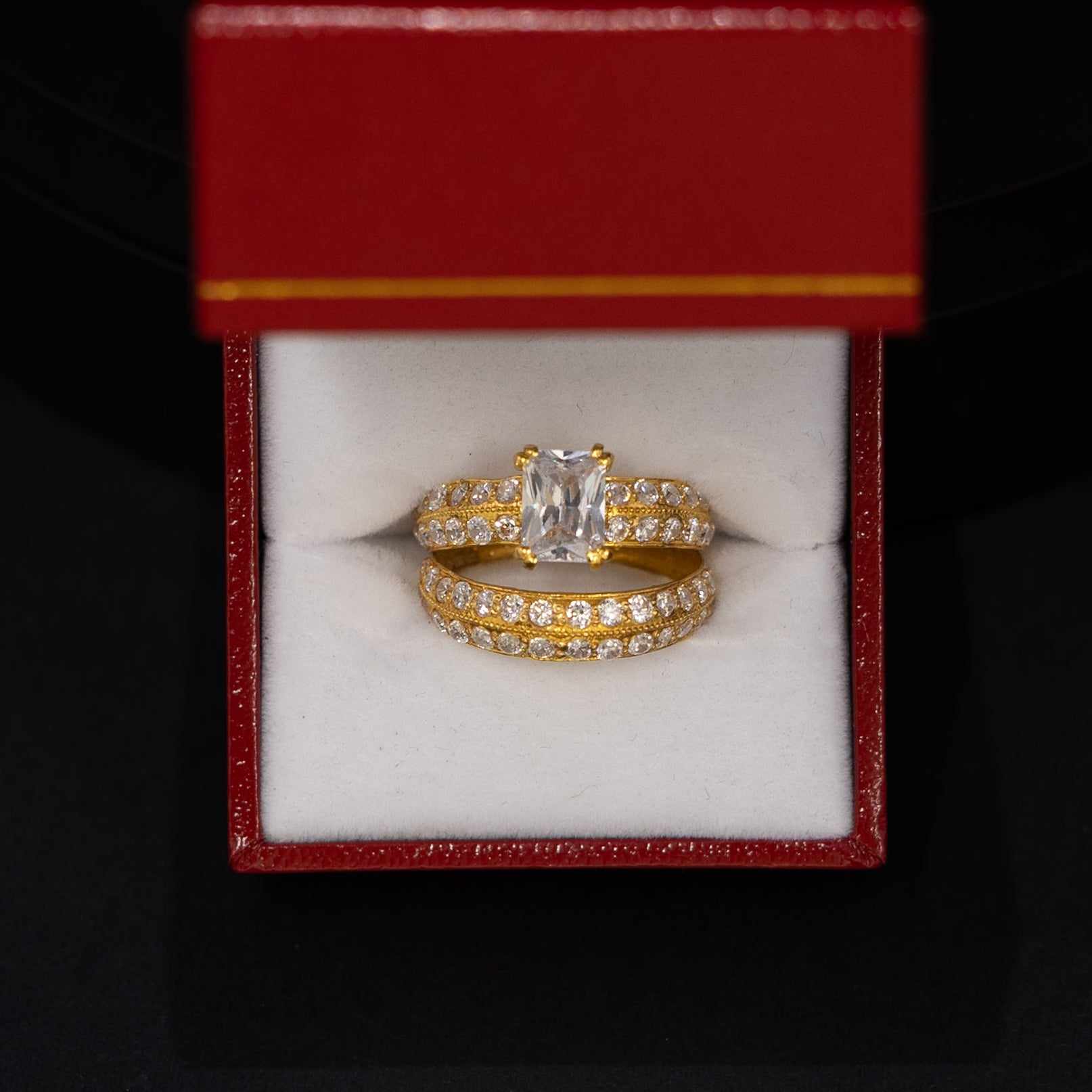 A yellow gold ring with a central clear gemstone displayed inside an open red jewelry box with a white interior, set against a dark black background.