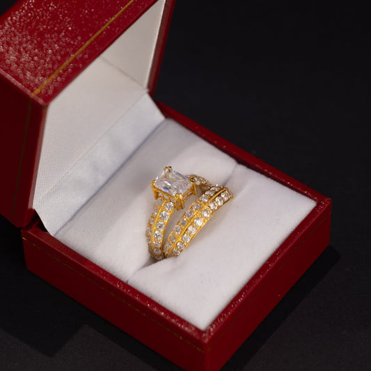 A gold ring with a clear rectangular gemstone and pavé-set details, displayed inside an open red jewelry box with a white velvet lining on a dark background.