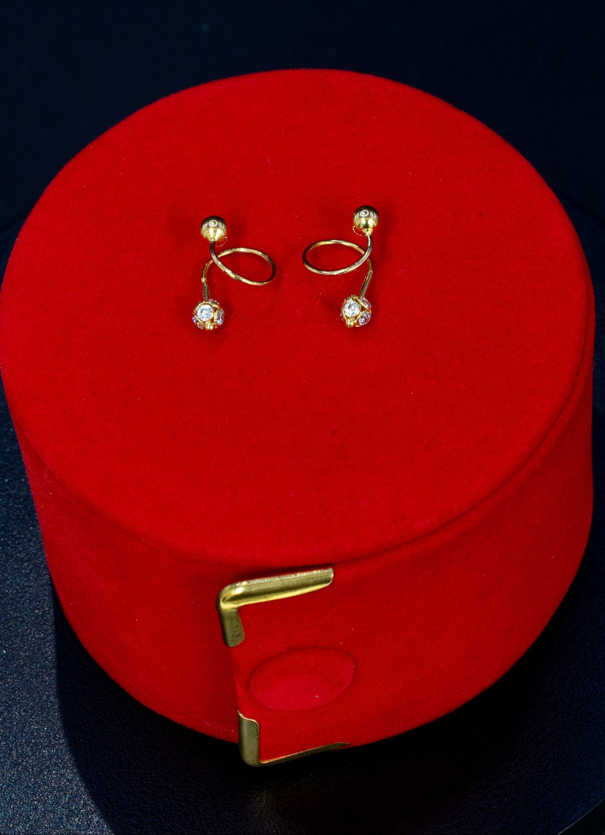 A pair of gold-toned hoop earrings with small clear gemstone drops, displayed on top of a round red jewelry box against a dark navy background.