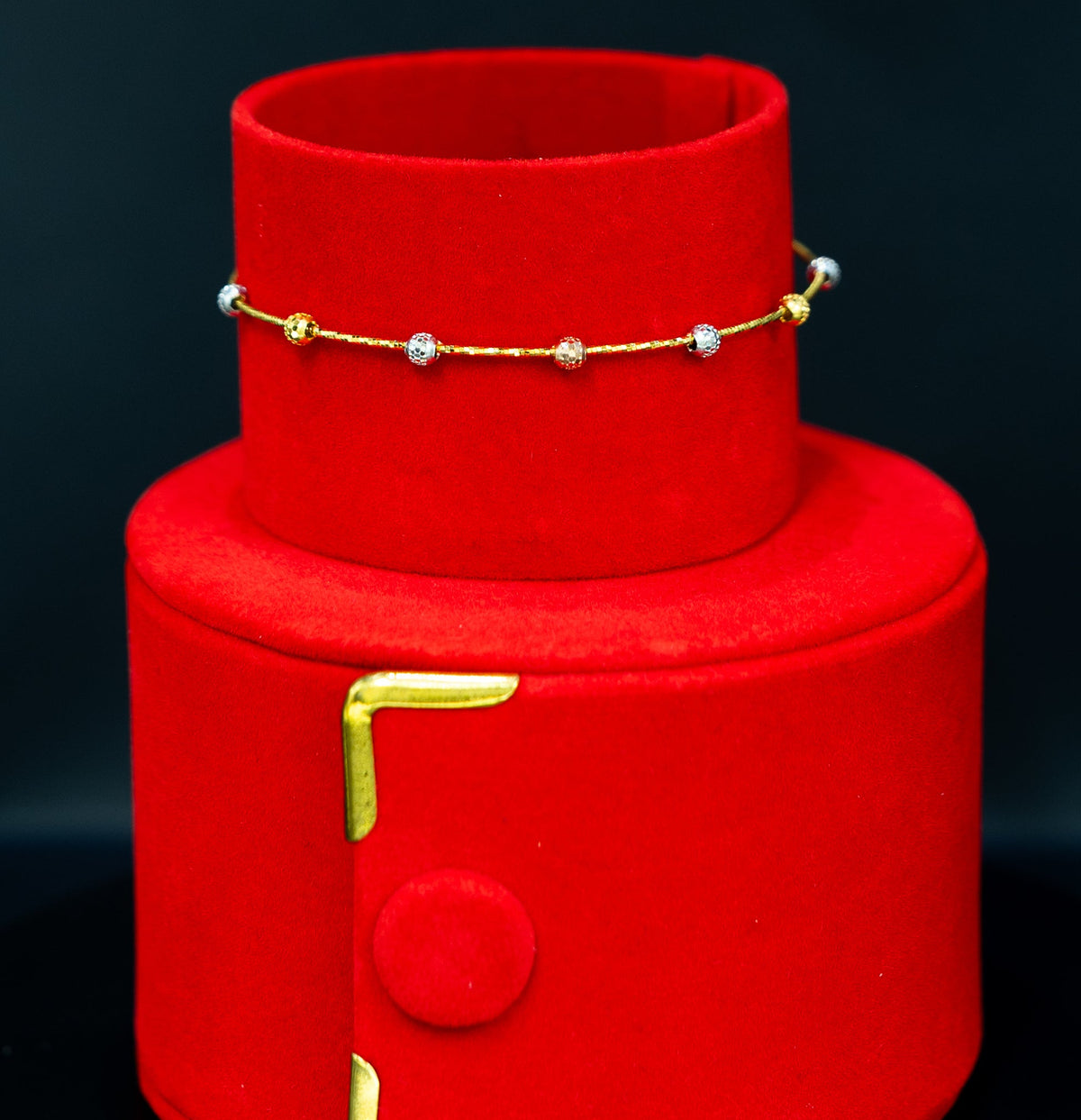 A gold chain bracelet with small white bead accents displayed on a red cylindrical jewelry stand against a dark background.