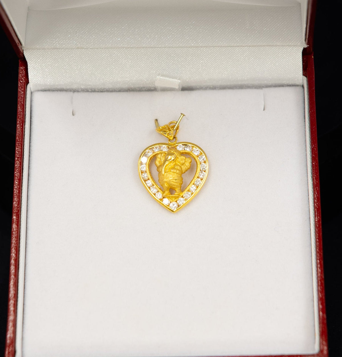 A gold heart-shaped pendant with a central figure, set with small clear stones around the border, displayed inside an open red jewelry box with a white insert.